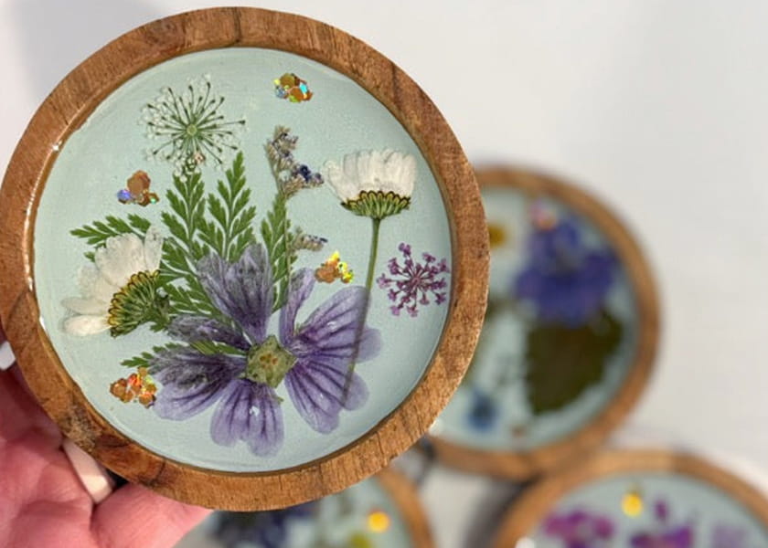 Hand holding a round wooden coaster filled with light blue resin, pressed purple and white flowers, and gold foil accents.