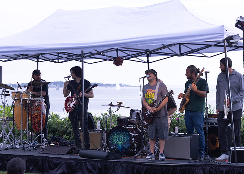A live musical band performing under a white canopy tent on an outdoor stage overlooking the ocean.