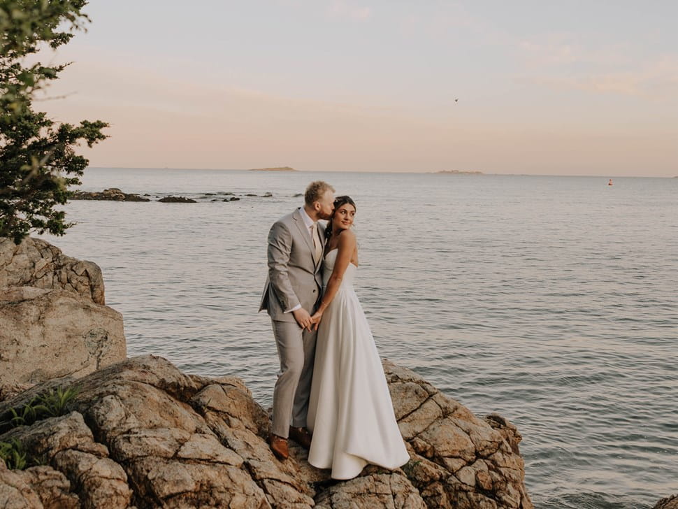 Groom in a light gray suit kissing bride's forehead on a rocky shore at ocean sunset.