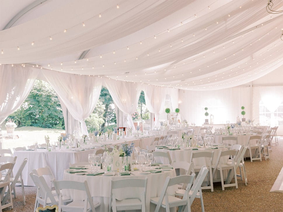 Wedding reception tent draped in white fabric and string lights with set dining tables.